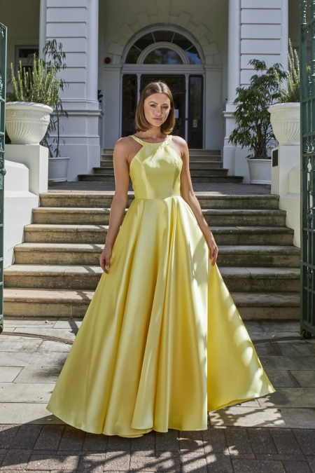 A woman stands outdoors in front of marble steps and an entrance with large white columns. She is wearing a flowing yellow satin gown with a halter neckline and a fitted bodice from the GLISS PO941 collection. Tall potted plants flank her on both sides at the top of the steps. Angels Formal Wear