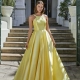 A woman stands outdoors in front of marble steps and an entrance with large white columns. She is wearing a flowing yellow satin gown with a halter neckline and a fitted bodice from the GLISS PO941 collection. Tall potted plants flank her on both sides at the top of the steps. Angels Formal Wear