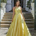 A woman stands on an outdoor staircase leading to a large building with tall, arched doorways. She is wearing a flowing, yellow satin gown with a halter neck design labeled GLISS PO941. Tall plants in large pots flank the stairway, and the sun casts shadows around her, highlighting her elegance. Angels Formal Wear