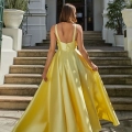 A woman in a flowing GLISS PO941 gown stands with her back facing the camera. The dress has a deep V back and she is gently holding the sides of her skirt. She is standing at the bottom of a stairway leading up to an entrance of a grand building with tall pillars. Angels Formal Wear