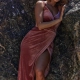 A woman wearing a sleeveless, velvet, rose-colored gown marked with the label HALEY TO889 leans against a textured rock wall. Her dress features a deep V-neck and a high slit that reveals her leg. She has short, wavy hair and is looking softly at the camera.
 Angels Formal Wear