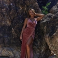 A woman poses confidently against a rocky backdrop. She is wearing a long, elegant, pinkish-brown velvet dress with a high slit, revealing her leg. Her right hand is resting behind her head, and she is looking towards the camera with a composed expression. The HALEY TO889 gown perfectly accentuates her poise. Angels Formal Wear