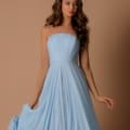 A woman with long, wavy brown hair is wearing a strapless, light blue HARLOW NBM1011 gown. She is standing against a beige background, gently holding one side of her dress. The HARLOW NBM1011 gown features vertical pleats on the bodice and flows elegantly down. Angels Formal Wear