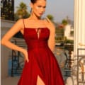 A woman poses outdoors in a sleeveless, red dress with a thigh-high slit, reminiscent of the ELOISE TW128 style. She has her hair styled in a bun and wears long ELOISE earrings. With one hand on her hip and the other near her face, she stands against a backdrop of decorative railing and blurred greenery. Angels Formal Wear