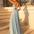 A woman stands on a set of stone steps, facing away from the camera. She wears a long, flowing light blue dress with a bow at the back, reminiscent of ELOISE TW128's timeless elegance, and her hair is styled in an elegant updo. Ornate black and gold railings decorate the background. Angels Formal Wear