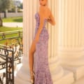 Hannah stands elegantly beside a white column, wearing a form-fitting, strapless lavender sequin gown with a high slit. She has her hair styled in an updo and is posing with one hand resting on the column. The background includes greenery and intricate black railings, enhancing the HANNAH TE307 elegance. Angels Formal Wear