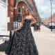 A woman stands on a city sidewalk wearing a strapless, floor-length black gown with intricate lace details, the kind of chic style you might find in an INDIE JX6067 fashion magazine. She poses with one hand holding the side of her dress. The backdrop features red brick buildings, a white door, and a street lamp. Angels Formal Wear