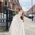 A woman in a beautiful lace strapless gown, possibly the INDIE JX6067 model, poses elegantly on a sidewalk in front of a brick building with black iron railings. The street exudes an indie charm, lined with similar buildings and parked cars under an overcast sky. Angels Formal Wear