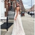 A woman in a white, strapless, lace gown stands on a sidewalk in an urban area with red brick buildings. She looks over her shoulder towards the camera, her long, wavy hair cascading down her back. The sky is partially cloudy, and a lamppost is visible in the background beside her INDIE JX6067. Angels Formal Wear