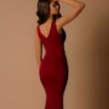 A woman stands sideways, her left hand touching the top of her head. She wears a KIMBRA NBM1010, a form-fitting, sleeveless, deep red formal dress with a V-shaped back. The background is a plain beige color, reminiscent of the sandy beaches of the Sunshine Coast. Angels Formal Wear