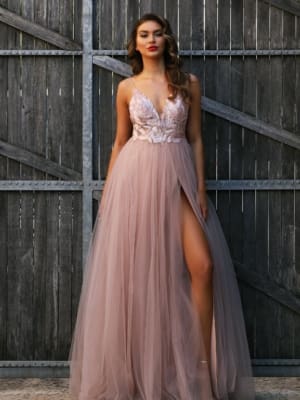 A woman stands in front of rustic wooden doors on the Sunshine Coast, wearing a flowing blush pink HEIDI JX3004 with a deep V-neckline, floral embroidery, and a high slit revealing one leg. Her hair is styled in loose waves, and she wears bold red lipstick, embodying the elegance of formal dresses. Angels Formal Wear