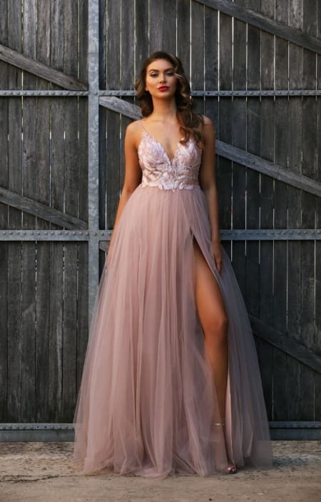 A woman stands in front of rustic wooden doors on the Sunshine Coast, wearing a flowing blush pink HEIDI JX3004 with a deep V-neckline, floral embroidery, and a high slit revealing one leg. Her hair is styled in loose waves, and she wears bold red lipstick, embodying the elegance of formal dresses. Angels Formal Wear