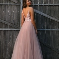 A woman faces a wooden fence while wearing a flowing, backless, light pink gown from HEIDI JX3004 on the Sunshine Coast. The strapped dress features lace details on the bodice and a long tulle skirt that adds an elegant and ethereal touch to her formal wear. Her hair is styled in soft waves. Angels Formal Wear