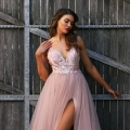 A woman stands against a rustic wooden gate, wearing a flowing light pink gown from HEIDI JX3004. With its deep V-neck and high slit, the formal wear beautifully complements her cascading waves as she gently touches her dress, looking off to the side. Angels Formal Wear