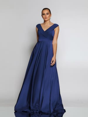 A woman stands against a neutral background wearing a sleeveless, deep blue evening gown with a V-neckline and a full, flowing skirt. Her hair is pulled back in a sleek updo, and she has a poised and elegant expression—perfect formal wear from JOELENE JX2116. Angels Formal Wear