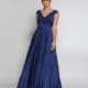 A woman stands against a neutral background wearing a sleeveless, deep blue evening gown with a V-neckline and a full, flowing skirt. Her hair is pulled back in a sleek updo, and she has a poised and elegant expression—perfect formal wear from JOELENE JX2116. Angels Formal Wear