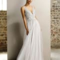 A woman in a white, sleeveless, V-neck gown with intricate beadwork stands against a backdrop of a brick wall and large window. Her dark hair is pulled back, and she has a graceful, elegant stance. The JORDI JX1077 flows gently to the floor, creating a classic look. Angels Formal Wear