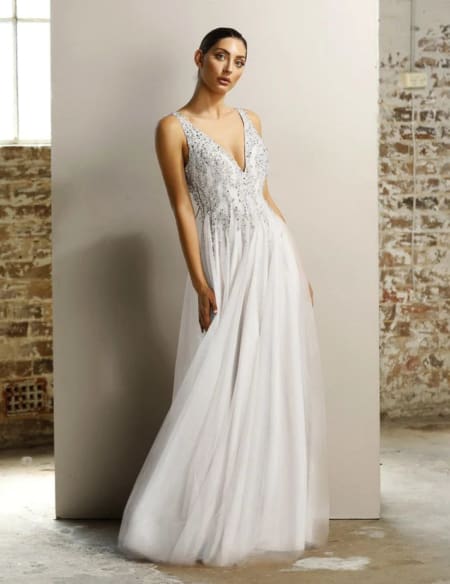 A woman in a white, sleeveless, V-neck gown with intricate beadwork stands against a backdrop of a brick wall and large window. Her dark hair is pulled back, and she has a graceful, elegant stance. The JORDI JX1077 flows gently to the floor, creating a classic look. Angels Formal Wear