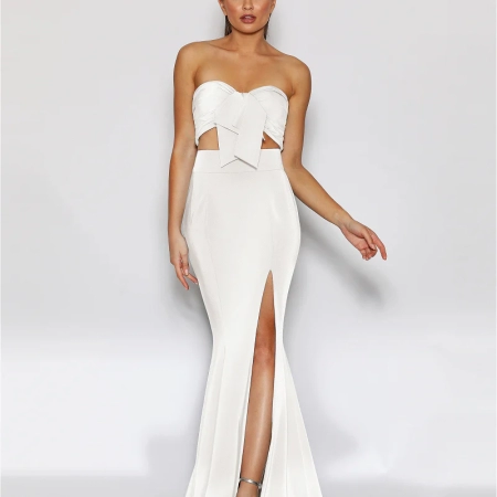 A woman is standing against a plain white background, wearing a strapless, white evening gown LIBERTY JX2043 with a large bow detail at the bust. The formal wear features a high slit on one side, revealing her left leg. She is wearing silver high-heeled sandals and her hair is styled up. Angels Formal Wear