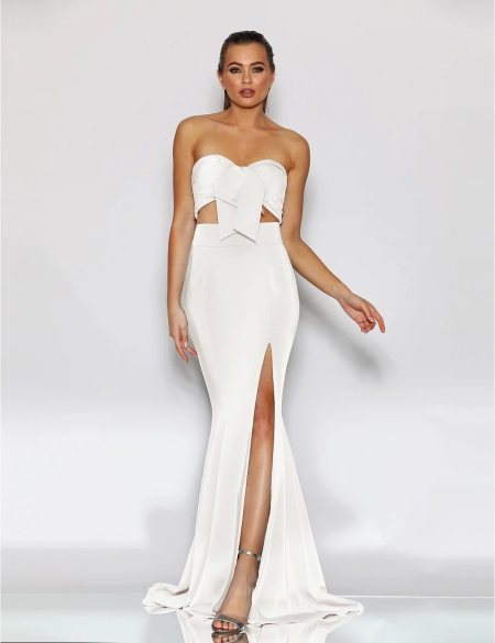 A woman is standing against a plain white background, wearing a strapless, white evening gown LIBERTY JX2043 with a large bow detail at the bust. The formal wear features a high slit on one side, revealing her left leg. She is wearing silver high-heeled sandals and her hair is styled up. Angels Formal Wear