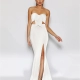 A woman is standing against a plain white background, wearing a strapless, white evening gown LIBERTY JX2043 with a large bow detail at the bust. The formal wear features a high slit on one side, revealing her left leg. She is wearing silver high-heeled sandals and her hair is styled up. Angels Formal Wear