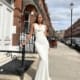 A woman stands elegantly in an off-white, form-fitting gown with a cowl neckline. She is on a city sidewalk, with tall, red-brick buildings and a black lamppost in the background. The sky is partly cloudy, adding soft light to the scene—a perfect look for any LILYLEE JX6080 shop specializing in formal wear. Angels Formal Wear