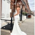 A woman stands on a city sidewalk wearing elegant formal wear—a form-fitting white gown with a long train. She looks over her shoulder at the camera. The background includes a brick building, white door, black iron fence, and a bicycle. The sky is partly cloudy, echoing the charm of LILYLEE JX6080. Angels Formal Wear