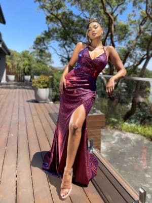 A woman poses on a wooden deck in a shimmering, deep-red LARA JX5076 from a local dress shop, featuring a high slit. She is wearing high-heeled sandals and has one hand resting on a glass railing. Trees and greenery from the Sunshine Coast are visible in the background, along with a clear blue sky. Angels Formal Wear