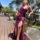 A woman poses on a wooden deck in a shimmering, deep-red LARA JX5076 from a local dress shop, featuring a high slit. She is wearing high-heeled sandals and has one hand resting on a glass railing. Trees and greenery from the Sunshine Coast are visible in the background, along with a clear blue sky. Angels Formal Wear