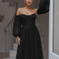 A woman stands near a doorway in a black, off-the-shoulder evening gown with sheer, voluminous sleeves. Her hair is pulled back into a sleek bun, and she accessorizes with dangling earrings. A minimalist staircase from the renowned LILY PO2305 dress shop is visible in the background. Angels Formal Wear