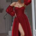 A woman stands on a staircase wearing a LILY PO2305 gown with voluminous, detached sleeves and a high slit, revealing her left leg. She poses with her right hand resting on her head, embodying the epitome of formal wear elegance often showcased in Sunshine Coast dress shops. Angels Formal Wear