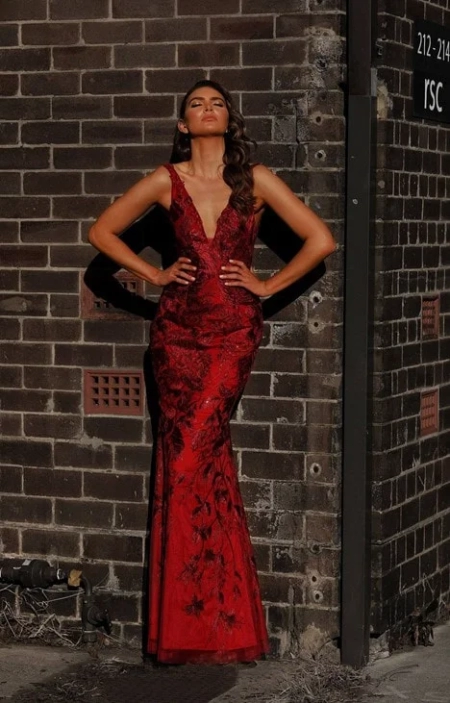 A woman in a red, sleeveless gown with a deep V neckline stands against a brick wall, embodying the elegance of formal wear. Her long, wavy hair cascades down as her hands rest confidently on her hips. The MARCELLE JX3045 from the Sunshine Coast's finest dress shop catches the light, creating strong shadows on the textured wall. Angels Formal Wear