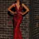 A woman in a red, sleeveless gown with a deep V neckline stands against a brick wall, embodying the elegance of formal wear. Her long, wavy hair cascades down as her hands rest confidently on her hips. The MARCELLE JX3045 from the Sunshine Coast's finest dress shop catches the light, creating strong shadows on the textured wall. Angels Formal Wear