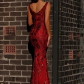 A woman wearing a red, sleeveless, full-length gown with floral details stands against a brick wall. She faces the wall with her back to the camera, showcasing the elegant design of the MARCELLE JX3045 dress. The wall has a sign reading "212-214 YSC. Angels Formal Wear