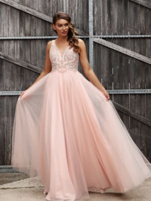 A woman stands in front of a wooden fence wearing a long, flowing, light pink MONTI JX3037 - SOLD from a Sunshine Coast dress shop. The formal wear features intricate beadwork on the bodice and a V-neckline. She holds out the skirt with both hands, showcasing the tulle layers. Her hair is styled in loose waves. Angels Formal Wear