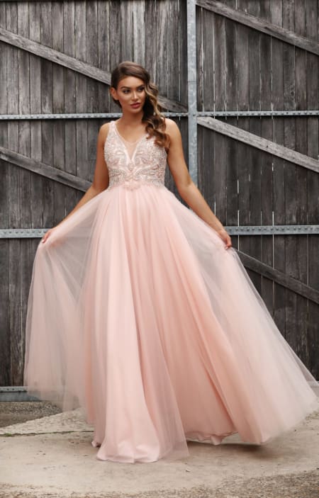 A woman stands in front of a wooden fence wearing a long, flowing, light pink MONTI JX3037 - SOLD from a Sunshine Coast dress shop. The formal wear features intricate beadwork on the bodice and a V-neckline. She holds out the skirt with both hands, showcasing the tulle layers. Her hair is styled in loose waves. Angels Formal Wear