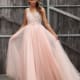 A woman stands in front of a wooden fence wearing a long, flowing, light pink MONTI JX3037 - SOLD from a Sunshine Coast dress shop. The formal wear features intricate beadwork on the bodice and a V-neckline. She holds out the skirt with both hands, showcasing the tulle layers. Her hair is styled in loose waves. Angels Formal Wear