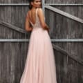 A woman stands in front of large wooden doors, facing slightly away from the camera. She is wearing a floor-length, light pink gown with a beaded bodice and sheer, flowing fabric from MONTI JX3037 - SOLD. Her hair is styled in loose waves, and she gazes over her shoulder. Angels Formal Wear