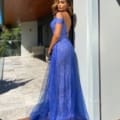 A woman stands on a patio on the Sunshine Coast, posing in a sparkly blue off-shoulder gown MATILDA JX5038 from a local dress shop. She looks over her shoulder towards the camera, one arm gracefully resting on the wall. The background features a wooden ceiling, glass railing, and lush greenery. Angels Formal Wear