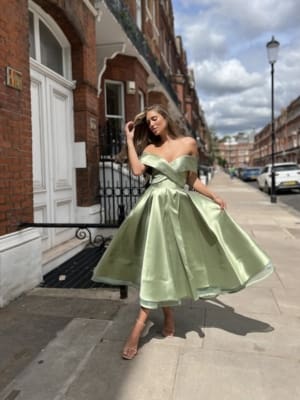 A woman stands on a residential street in front of a brick building, wearing an off-the-shoulder, light green MARILYN JX6026 with a full skirt. She has one hand raised to her head, adjusting her hair, as the Sunshine Coast sun casts shadows on the sidewalk. Angels Formal Wear