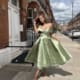 A woman stands on a residential street in front of a brick building, wearing an off-the-shoulder, light green MARILYN JX6026 with a full skirt. She has one hand raised to her head, adjusting her hair, as the Sunshine Coast sun casts shadows on the sidewalk. Angels Formal Wear