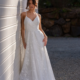 A woman in a white lace wedding dress with thin straps, labeled BRIONY PO972, leans against a white wall. Her hair is styled in an updo as she stands on a gravel path surrounded by greenery and rocks. The sunlight casts a soft shadow on the wall. Angels Formal Wear