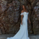 A woman stands on a sandy beach wearing a fitted white wedding dress with off-the-shoulder sleeves and a flowing train. Behind her, the rocky cliff face, resembling the rugged textures of DAISY TC379, displays various hues. The woman has wavy hair and gazes to the side. Angels Formal Wear