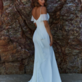 A person in an elegant, off-the-shoulder white dress stands in front of a textured rocky background. The dress features ruffled details and a long, flowing train reminiscent of the grace found in DAISY TC379. The individual has wavy hair and their back is slightly turned towards the camera. Angels Formal Wear