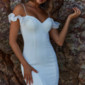 A woman in a fitted white off-the-shoulder dress with ruffled sleeves stands against a textured rock wall adorned with DAISY TC379 blooms. She has wavy, shoulder-length hair and is holding a delicate hand up beside her face while looking into the camera with a serene expression. Angels Formal Wear