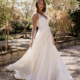 A woman wearing ESTELLE TC399, a white sleeveless wedding dress with lace detailing on the bodice, stands outdoors on a gravel path. She is holding the sides of her dress, which fan out around her. Behind her are various green plants and trees. Angels Formal Wear