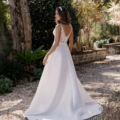 A bride wearing a white wedding dress with a lace bodice and a long, flowing skirt stands on a gravel pathway in a garden. She faces away from the camera, glancing over her shoulder, with greenery and sunlight filtering through the trees around her. This elegant scene captures the timeless charm of ESTELLE TC399. Angels Formal Wear