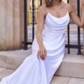 A woman in a white satin gown with thin straps leans against a stone step in an outdoor setting. She has shoulder-length hair and is wearing dangling earrings. In the background, there is a black wrought-iron fence and a stone building marked ESTHER PO944. Angels Formal Wear