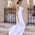 A woman in a flowing white gown, reminiscent of ESTHER PO944, walks down stone steps outside, flanked by tall, ornate columns. She has her hair tied back and is wearing pearl earrings. The background includes a black iron fence and a light-colored building with arched windows labeled PO944. Angels Formal Wear