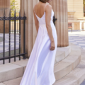 A woman stands outdoors on stone steps wearing a flowing, sleeveless white dress. She faces away from the camera, highlighting the open back and elegant drape of the ESTHER PO944. Tall, beige stone columns and a wrought iron fence are visible in the background. Angels Formal Wear